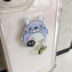 May include: A clear phone case with a blue and white Totoro character charm attached. The charm has glitter and a small black and white character with large eyes hanging from it.