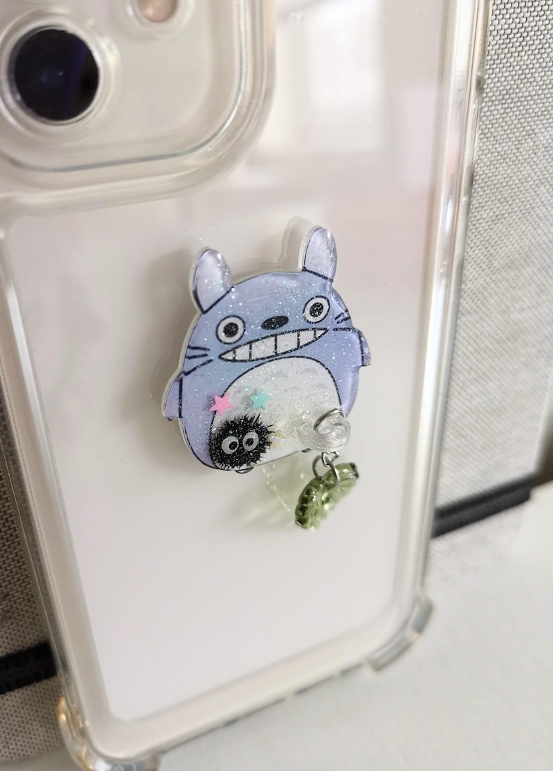 May include: A clear phone case with a blue and white Totoro character charm attached. The charm has glitter and a small black and white character with large eyes hanging from it.