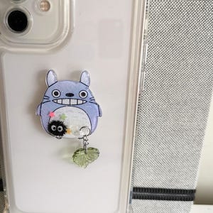 May include: A clear phone case with a blue and white Totoro character charm attached. The charm has a small black and white character hanging from it with a green leaf charm.