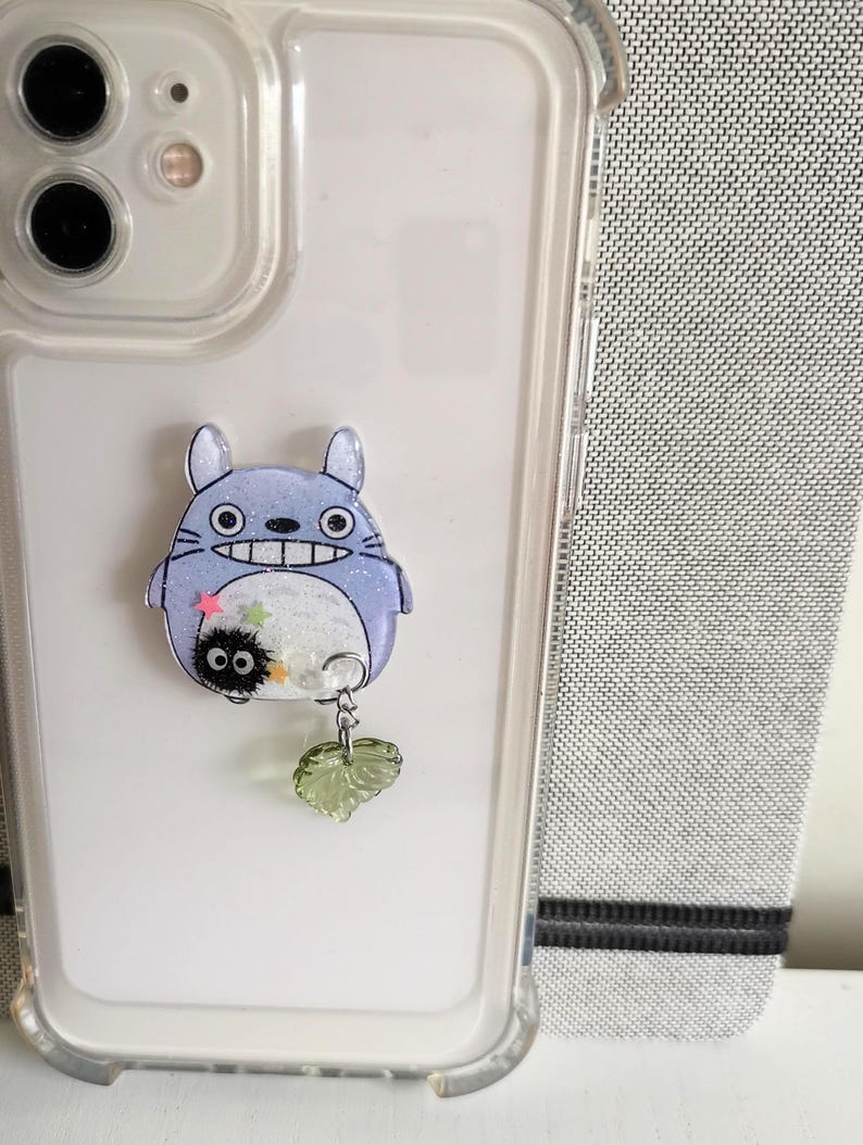 May include: A clear phone case with a blue and white Totoro character charm attached. The charm has a small black and white character hanging from it with a green leaf charm.