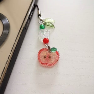 May include: A beaded phone charm featuring a red apple pendant with a green leaf and a silver-colored metal clasp. The charm includes clear flower beads, green and red beads, and is attached to a phone.