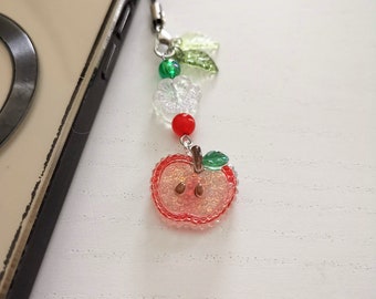 May include: A beaded phone charm featuring a red apple pendant with a green leaf and a silver-colored metal clasp. The charm includes clear flower beads, green and red beads, and is attached to a phone.