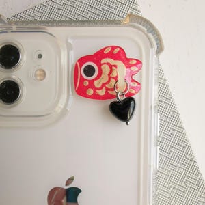 May include: A clear phone case with a red and gold fish charm attached to a black heart charm. The phone case has a white background and an Apple logo.