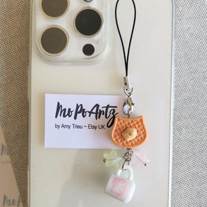 May include: A phone charm with a black strap and silver-tone hardware. The charm features a cat-shaped waffle, star-shaped charms, and a white mug with a pink design. The text "Mo Po Artz" is visible on a white label.
