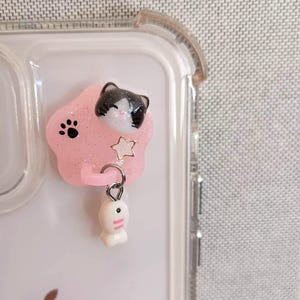 May include: A clear phone case with a pink paw-shaped charm. The charm features a black and white cat face, a star, and a dangling white fish. The charm is attached to the phone case with a silver ring.