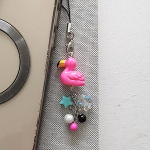 May include: A pink flamingo charm with a yellow beak, hanging from a silver-colored clasp. Below the flamingo are a turquoise star, a clear charm, and several beads in white, black, and pink. The charm is attached to a black cord.