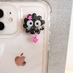 May include: A clear phone case with a black and glitter character charm attached. The charm has a pink star dangling from it.