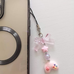 Könnte beinhalten: Ein rosa-weißer Handy-Anhänger mit einer rosa-weißen Schleife, einem Stern und einem rosa-weißen Gamecontroller. Der Anhänger ist an einem schwarzen Handyband befestigt.