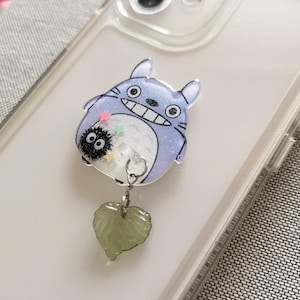 May include: A clear phone case with a Totoro character charm attached. The charm is made of resin and has glitter embedded in it. The charm is attached to a green heart-shaped charm.