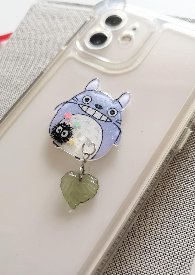 May include: A clear phone case with a Totoro character charm attached. The charm is made of resin and has glitter embedded in it. The charm is attached to a green heart-shaped charm.