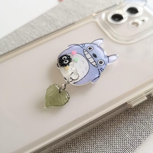 May include: A clear phone case with a blue and white character from the movie My Neighbor Totoro. The character is holding a small black character with white eyes. A green leaf charm hangs from the character.