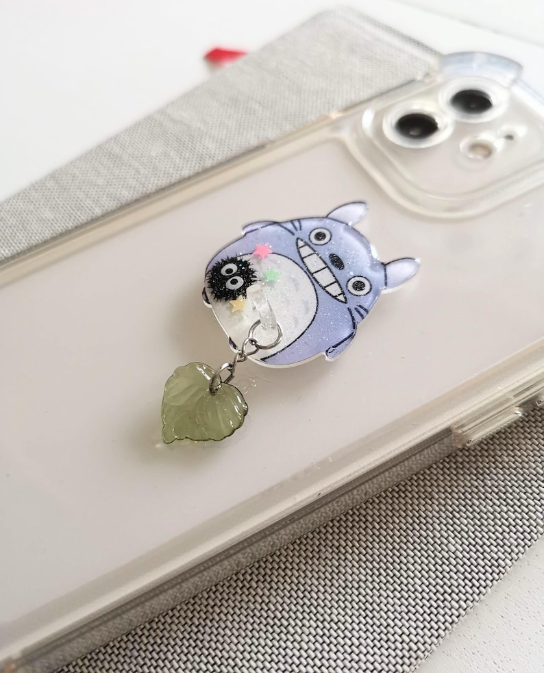 May include: A clear phone case with a blue and white character from the movie My Neighbor Totoro. The character is holding a small black character with white eyes. A green leaf charm hangs from the character.