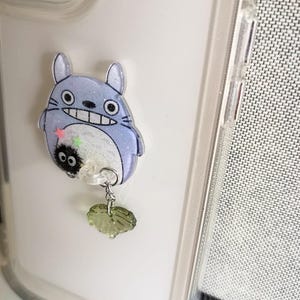 May include: A clear phone case with a blue and white Totoro character charm attached. The charm has a small black and white character hanging from it with a green leaf charm.