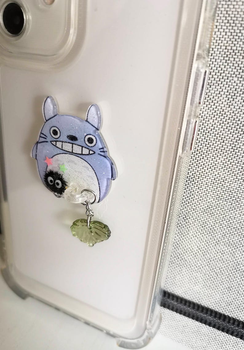 May include: A clear phone case with a blue and white Totoro character charm attached. The charm has a small black and white character hanging from it with a green leaf charm.
