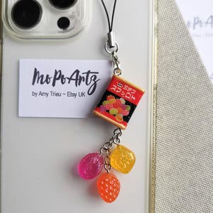 May include: A phone charm with a miniature candy bag and colourful gummy sweets. The bag has Japanese text and a red and black design. The charm includes pink, orange, and yellow sweets. The phone case is clear.