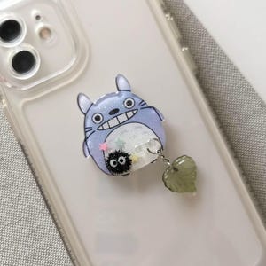 May include: A clear phone case with a blue and white Totoro character phone grip with a small black Totoro charm attached to a green heart charm.