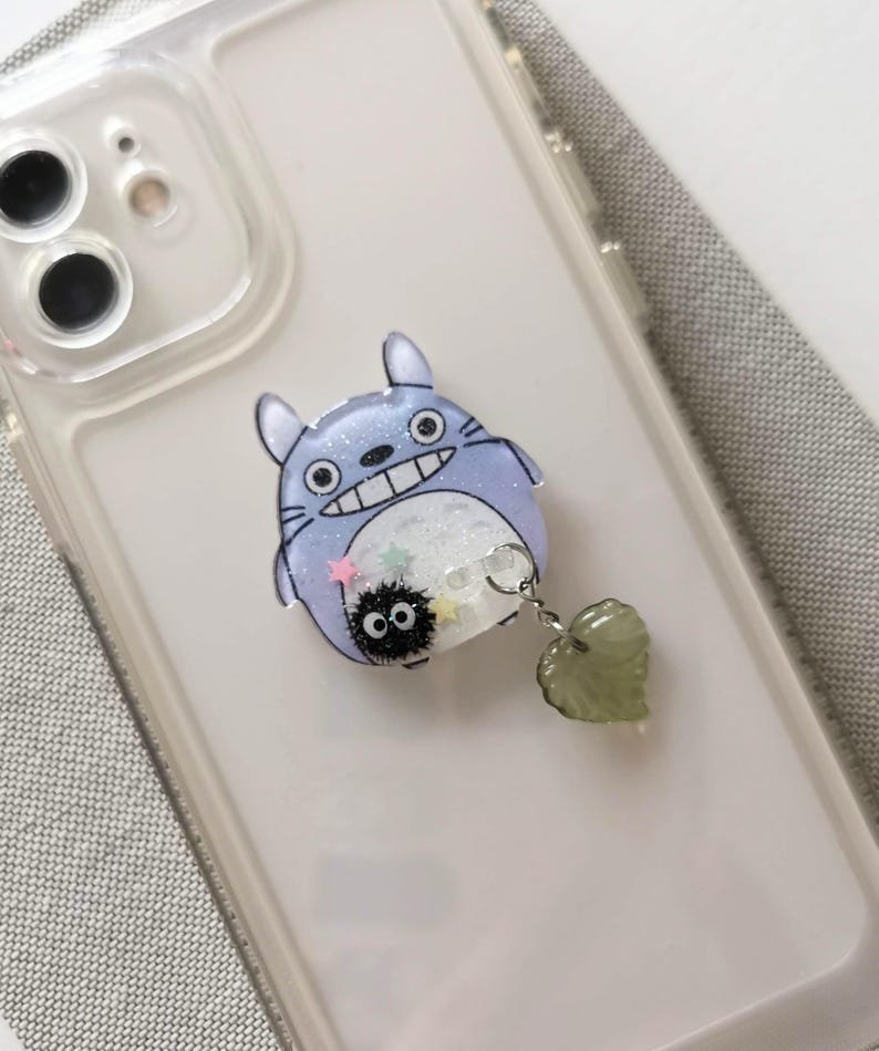 May include: A clear phone case with a blue and white Totoro character phone grip with a small black Totoro charm attached to a green heart charm.