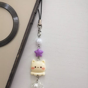 Op de afbeelding: Een crèmekleurige kattenbedel met roze wangen en een kleine mond, hangend aan een telefoon. De bedel bevat een paarse ster, witte kralen en een doorzichtige blad-vormige bedel met een parel. De bedel is bevestigd aan een zwart koord.