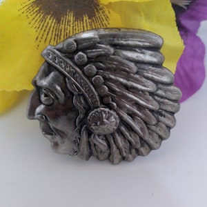 May include: A silver-toned metal pin depicting a Native American chief wearing a headdress with feathers. The pin is detailed and has a realistic look.