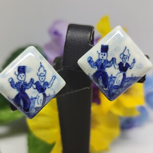 Könnte beinhalten: Ein Paar blaue und weiße Keramikohrringe mit einem holländischen Paar in traditioneller Kleidung, das tanzt. Die Ohrringe sind diamantförmig.