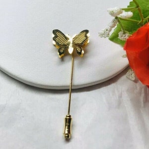 Könnte beinhalten: Eine goldfarbene Schmetterlingsbrosche mit strukturiertem Design. Die Brosche ist an einer langen, dünnen Nadel befestigt.