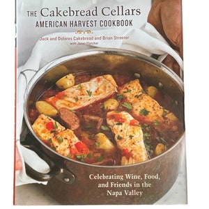May include: The Cakebread Cellars American Harvest Cookbook, a book with a red background and a photo of a pot of food with fish, potatoes, and peppers. The text on the cover reads "Celebrating Wine, Food, and Friends in the Napa Valley."