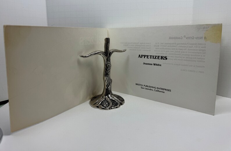 May include: A silver metal tree-shaped stand for holding napkins or other small items. The stand is on a white surface with a book titled "Appetizers" by Joanna White. The book is published by Bristol Publishing Enterprises in San Leandro, California.