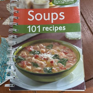 May include: A spiral-bound cookbook titled "Soups 101 recipes" with a photo of a bowl of soup on the cover. The cover has a red and green banner with the title. The soup in the bowl appears to have chicken, spinach, and tomatoes.