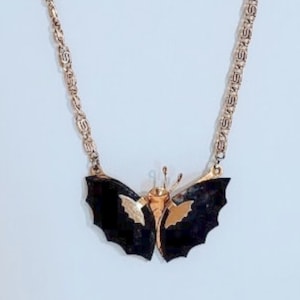 Puede incluir: Un collar de cadena dorada con un colgante de mariposa negra. La mariposa tiene detalles dorados en las alas.