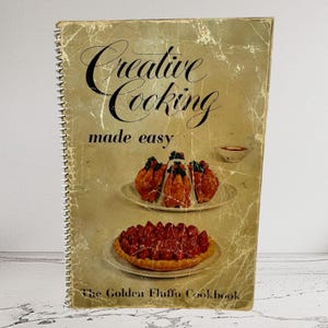 Puede incluir: Libro de cocina vintage con una cubierta amarilla y el título "Creative Cooking made easy" en letras cursivas negras. La cubierta presenta una foto de un plato de pollo y una tarta de fresa. El libro de cocina se titula "The Golden Fluffo Cookbook".
