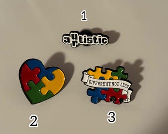 May include: Three enamel pins with autism awareness themes. One pin reads "autistic" in black and white. Another is a heart-shaped puzzle. The third pin is a puzzle with a banner that reads "Different Not Less."