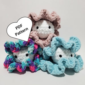 May include: Three crocheted plush toys shaped like a clam with a white face and black eyes. The clams are in pink, blue, and a mix of pink, blue, and purple. A white heart with the text "PDF Pattern" is in the background.