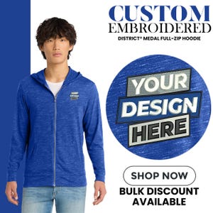 May include: A blue full-zip hoodie with the text "YOUR DESIGN HERE" on the chest and a larger graphic with the same text. The hoodie is part of the DISTRICT® MEDAL line and is custom embroidered. The image also includes the text "SHOP NOW" and "BULK DISCOUNT AVAILABLE."