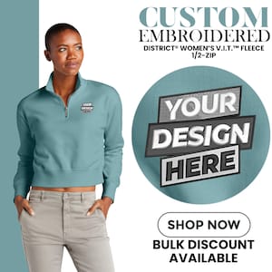 May include: A teal District Women's V.I.T. fleece 1/2-zip sweatshirt with a custom embroidered design. The sweatshirt has a zipper and a small embroidered logo. The image also includes text that says "Your Design Here" and "Shop Now".