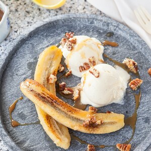 May include: Two scoops of vanilla ice cream topped with chopped pecans and drizzled with caramel sauce, served over two slices of caramelized bananas on a gray plate.