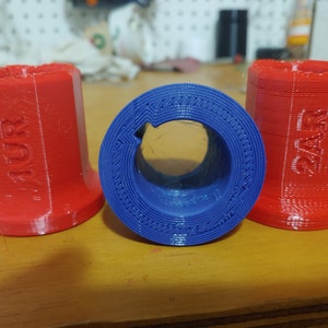 Può includere: Tre oggetti stampati in 3D, due rossi e uno blu. Gli oggetti rossi sono cilindrici con il testo "11R" e "22F" stampato su di essi. L'oggetto blu è un anello con un foro al centro.