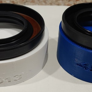 Könnte beinhalten: Zwei runde, schwarze Gummidichtungen mit einem weißen und blauen Kunststoffsockel. Der weiße Sockel trägt den Aufdruck "547013". Der blaue Sockel trägt den Aufdruck "4R Runner".
