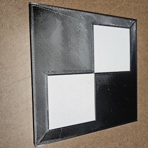 May include: A square, flat object featuring a high-contrast checkerboard pattern. The design consists of two solid white squares positioned diagonally against a black background, all framed by a thick, textured black border with visible lines.