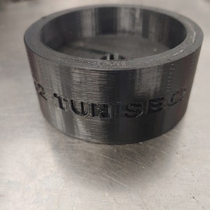 Könnte beinhalten: Ein schwarzer 3D-gedruckter Zylinder mit dem Text "2 TUNISED" auf der Seite.