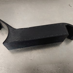 May include: A black, 3D-printed object with a textured surface. The design features a rectangular base with a curved section on one end and a raised, angled section on the other. The object appears to be made of plastic.
