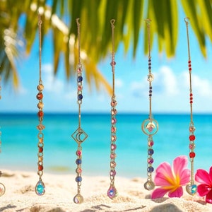 May include: Several decorative suncatchers with gold chains and colorful beads hang against a beach backdrop. Each suncatcher features a unique design, including crystals, geometric shapes, and various bead arrangements. Pink flowers are in the foreground.