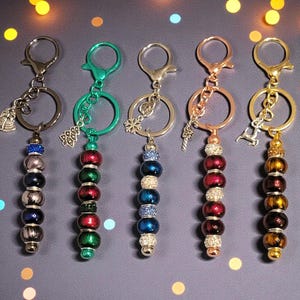 May include: A collection of five keychains with decorative beaded pendants. Each keychain features a metal ring and clasp, with pendants in various colors including silver, green, red, and gold. Charms include a bell, a tree, a snowflake, a key, and a reindeer.