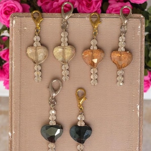 May include: Five heart-shaped crystal charms on a white background. Each charm is attached to a silver or gold chain with a clasp. The charms are in different colors: clear, light gold, light brown, and dark gray.