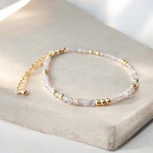 May include: A delicate bracelet with small, pale beads and gold-coloured accents. The bracelet has a gold-coloured chain and clasp. The jewellery is displayed on a light-coloured surface.