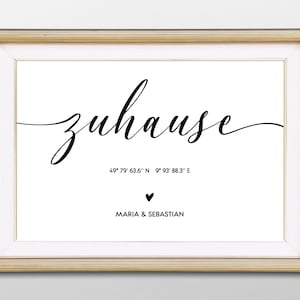 Puede incluir: Un cuadro enmarcado con la palabra alemana "zuhause" en escritura cursiva. Debajo, coordenadas y los nombres "Maria & Sebastian" con un símbolo de corazón. El marco es dorado.