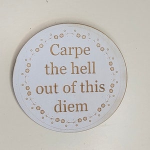 May include: A circular, white wooden sign featuring the engraved text "Carpe the hell out of this diem" in a brown font. The sign is encircled by a floral pattern. The background is a light beige colour.