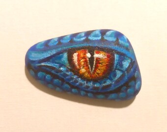 May include: A hand-painted rock with a blue and black patterned design and a single orange eye with a white pupil.