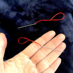 May include: Two needles with red thread, one needle is threaded and the other is not.