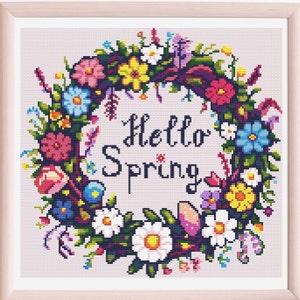 Puede incluir: Un diseño de bordado de punto de cruz con una corona floral colorida con el texto "Hello Spring" en el centro.