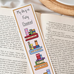 May include: A pink and white bookmark with a gold border. The bookmark features the text "My day is Fully Booked" and three stacks of colorful books.